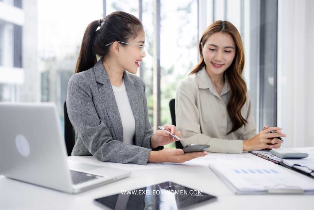 Two women sitting at a desk in a bright office, smiling and discussing work.